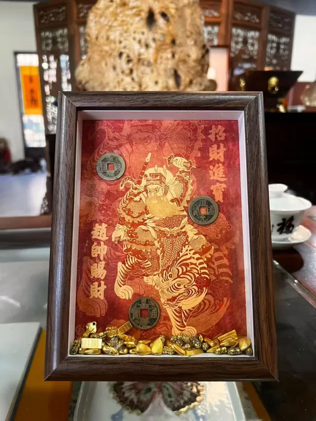 A framed red-and-gold prosperity image with a figure on a tiger and coin-like discs.