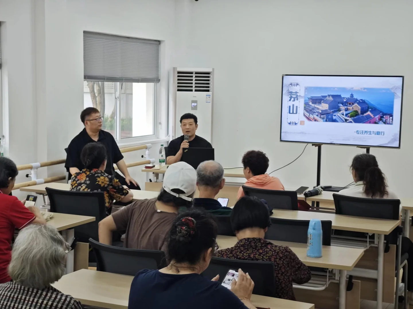 Participants seated in a classroom listening to two speakers beside a screen showing Maoshan